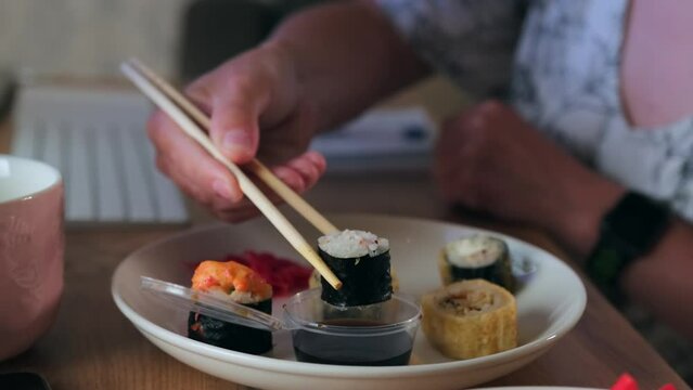 Female hands in Chinese chopsticks dip rolls in soy sauce, young girl freelancer in glasses is having dinner while working in her home office appetizingly eating smacking and watching movie on monitor