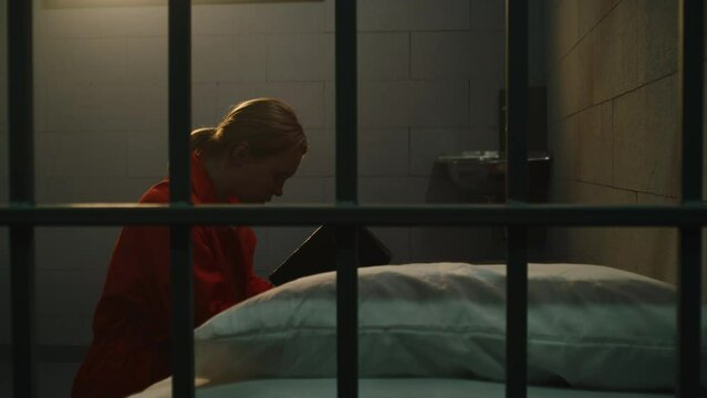 Female prisoner in orange uniform kneels near the bed, prays to God in prison cell holding Bible. Woman criminal serves imprisonment term for crime in jail, detention center or correctional facility.