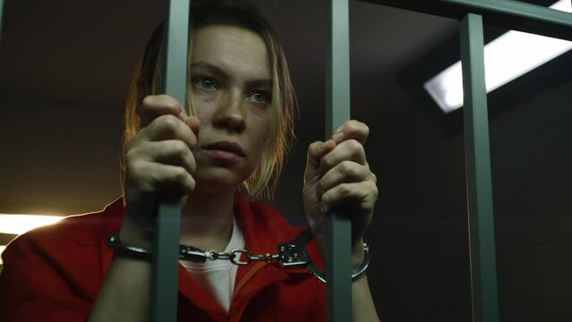 Shocked female prisoner in orange uniform holds metal bars, stands in prison cell in handcuffs. Woman criminal serves imprisonment term in jail. Depressed inmate in correctional facility. Portrait.