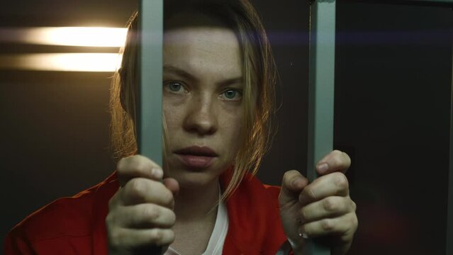 Scared female prisoner in orange uniform holds metal bars, stands in prison cell in handcuffs, looks at camera. Woman criminal serves imprisonment term for crime in jail or detention center. Portrait.