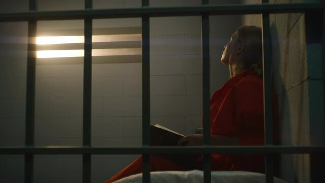 Female prisoner in orange uniform sits on bed behind metal bars, reads Bible in prison cell, looks at barred window. Woman criminal serves imprisonment term for crime in jail or correctional facility.