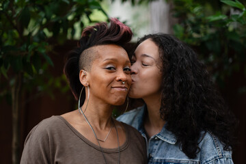 Two women express their love. One of them kisses the other one on the cheek. They are a lesbian couple, and Black and multiracial, expressing additional backstory.