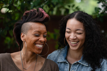 A lesbian couple laughs joyfully, showing a moment of love, togetherness, and happiness as Black, multiracial, and LGBTQ women. They are outdoors, in a garden or park setting.