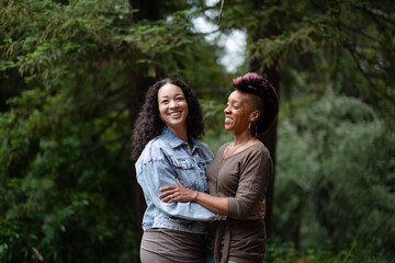 Naklejka premium With big smiles as they celebrate their engagement, a multiracial lesbian couple poses for portraits in a forest. They are facing each other and in a loving embrace. 
