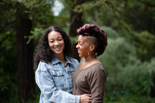 With big smiles as they celebrate their engagement, a multiracial lesbian couple poses for portraits in a forest. They are facing each other and in a loving embrace. 