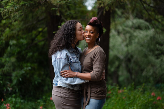 A lesbian couple celebrate their engagement with joyful and romantic photos in a backyard. They share a kiss, surrounded by the rich green foliage of pines and oaks. 