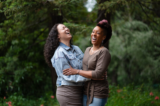 Holding each other in a romantic embrace, a lesbian couple has a joyful moment of laughter together. One has a pink mohawk, and the other has long, curly, natural hair. They are surrounded by trees.