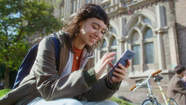 Young happy girl sitting outdoors on college campus, texting on mobile phone and smiling. Zoom shot, low angle view