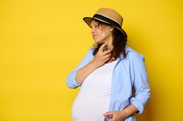 Beautiful pregnant woman in blue casual shirt and straw hat, applies perfume on her neck, isolated on yellow background.