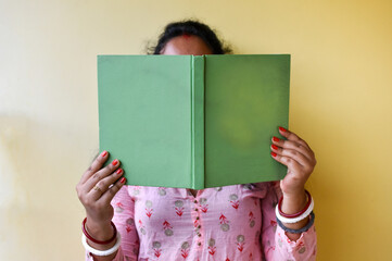 An Indian woman reading a book and covering her face with a yellow background. Selective focus on the book.
