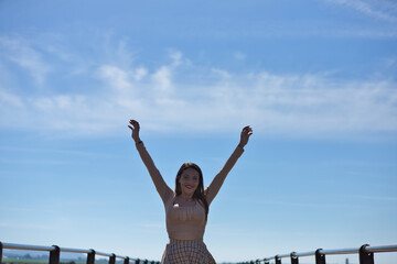 Pretty young woman with long brown hair wearing a short skirt and shirt raises her arms happily as she crosses the wooden bridge. In the background the blue sky and white clouds. The woman is freedom.