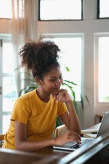 Portrait of young African American woman sitting in cozy home working remotely, surfing internet on laptop