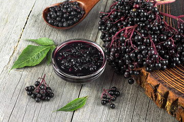 Delicious homemade black elderberry syrup in glass jar and bunches of black elderberry with green leaves on wooden desk. - obrazy, fototapety, plakaty