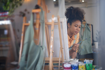 Charming female artist sitting in front of an easel and dipping brush into color palette