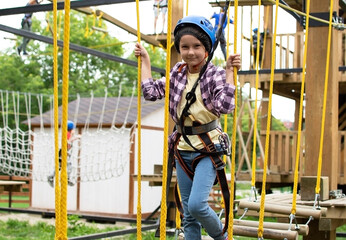 Happy child in helmet and protective gear enjoys classes in climbing adventure park on summer day. Kid is climbing on a rope playground