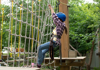 Happy child in helmet and protective gear enjoys classes in climbing adventure park on summer day. Kid is climbing on a rope playground