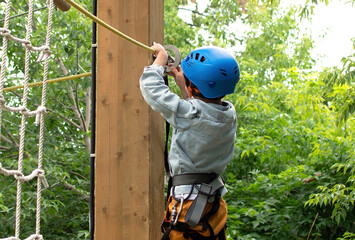 Happy child in helmet and protective gear enjoys classes in climbing adventure park on summer day....