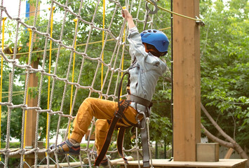 Happy child in helmet and protective gear enjoys classes in climbing adventure park on summer day. Kid is climbing on a rope playground