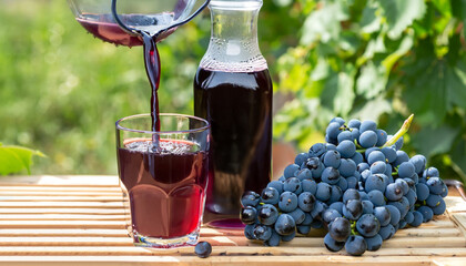 fresh grape juice pouring into glass from jug with ripe bunch of dark blue berries on crate as...