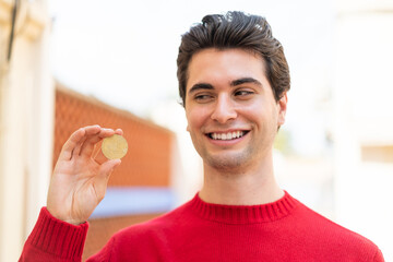 Young handsome man holding a Bitcoin at outdoors with happy expression