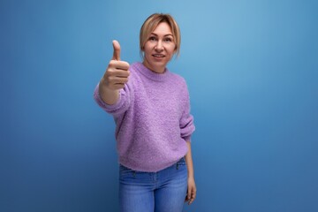 portrait of pleasant charming pleasant blond young woman in purple sweater with like ok gesture on blue background with copy space