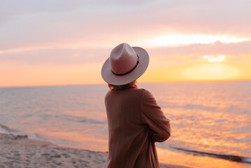 Lonely pensive woman in coat and hat looking at sunset, enjoying sea. Rear view of an...