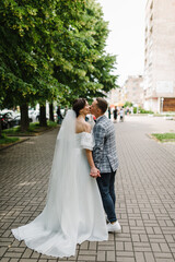 Fototapeta na wymiar Kiss. Young, stylish groom and beautiful bride in white dress walking in the alley in the city against the background of buildings, trees. Wedding photography of newlyweds on the street. Back view.