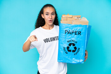 Young woman holding a recycling bag full of paper to recycle isolated on blue background surprised and pointing front