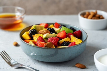 Healthy fruit salad in a grey bowl made with avocado, strawberry, raspberry, orange, blackberry, almonds and peaches. Yogurt, honey behind. Grey background