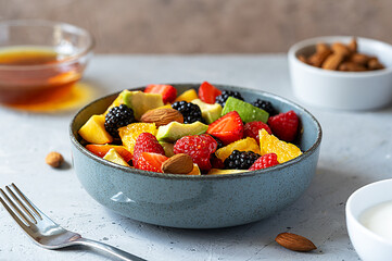Healthy fruit salad in a grey bowl made with avocado, strawberry, raspberry, orange, blackberry, almonds and peaches. Yogurt, honey behind. Grey background