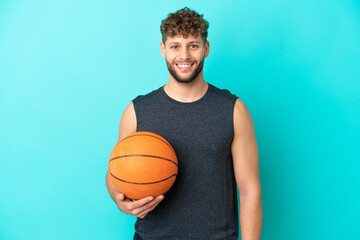 Handsome young man playing basketball isolated on blue background posing with arms at hip and smiling