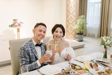 Cheers. Newlyweds holding glasses of champagne to toast at table in banquet hall restaurant. Bride...