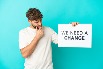 Young handsome caucasian man isolated on blue background holding a placard with text We Need a...