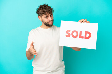 Young handsome caucasian man isolated on blue background holding a placard with text SOLD making a...