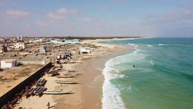A sandy beach of turquoise waters in the fishing village of Kayar (Senegal). The aerial view advances as the waves reach the shore, which is peppered with a few colorful traditional fishing boats.