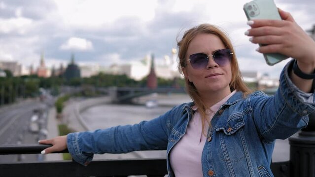 A young woman in sunglasses takes a selfie standing on a bridge overlooking the city and the river.