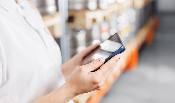 Factory worker with computer tablet inspecting kegs with beer in warehouse of modern brewery stock, blurred background