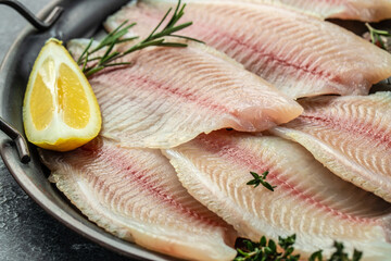 raw Tilapia fish, skinless meat with lemon and spices on a dark background.