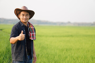 Naklejka na ściany i meble Portrait of happy Asian Thai male farmer wears cowboy hat and shows thumb up for good product at his green organic rice paddy field. Concept agricultural occupation. Satisfied in agricultural product.