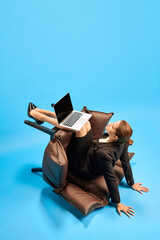 Serious business woman, worker sitting on fallen chair and working on laptop on her knees against blue studio background. Concept of business, working routine, deadlines, freelance, office, ad