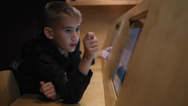 Two children are sitting in the library in front of an electronic tablet with a touch screen while studying. A boy reads an interesting book or article in a modern library from a tablet on the wall.
