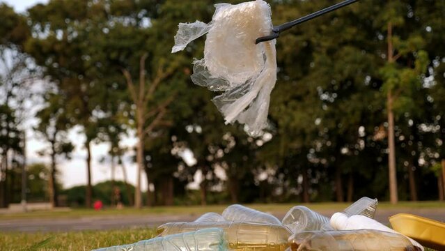 Close-up of a female activist collecting plastic garbage with tongs on the green grass. Volunteers help to save nature from plastic waste pollution.