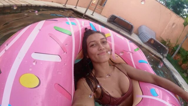 Beautiful girl in a swimsuit floats in a pink inflatable circle in the shape of a donut. Cheerful female smiling through donut inflatable ring. Woman relaxing on donut lilo in the pool water in hot