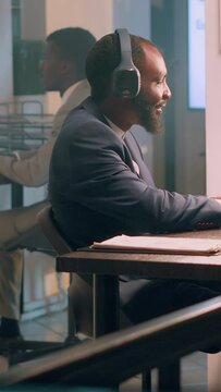 Vertical video African american worker enjoying music to take his mind away from boring routine project tasks during nightshift. Businessman in office with headphones on comparing marketing graphs and