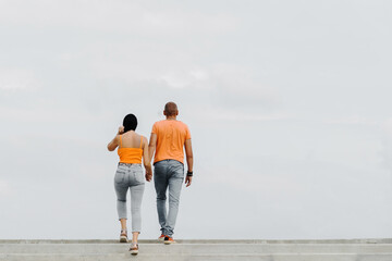 A young couple in orange t-shirts walk holding hands. The guy and the girl climb the stairs against the backdrop of a cloudy sky. Back view. Unrecognizable person. Space