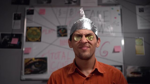 Strange stupid young man conspiracy theorist in protective foil cap with bitcoin coins in his eyes looking at camera. Conspiracy theory concept. Fake and alternative facts.