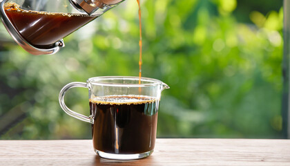 closeup coffee pouring from french press into glass cup on wooden table with green blurred...