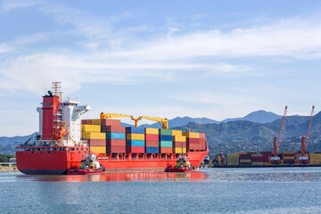 A large red cargo ship loaded with multicolored shipping containers enters the port to the...