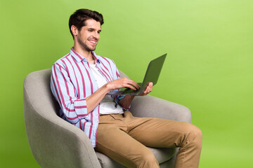Portrait of optimistic clever person wear stylish clothes sit in armchair look at laptop typing post isolated on green color background