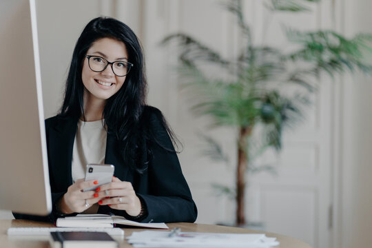 Cheerful office worker, stylish in black jacket, uses smartphone happily while working on an important project in a modern coworking space. Modern technologies.
